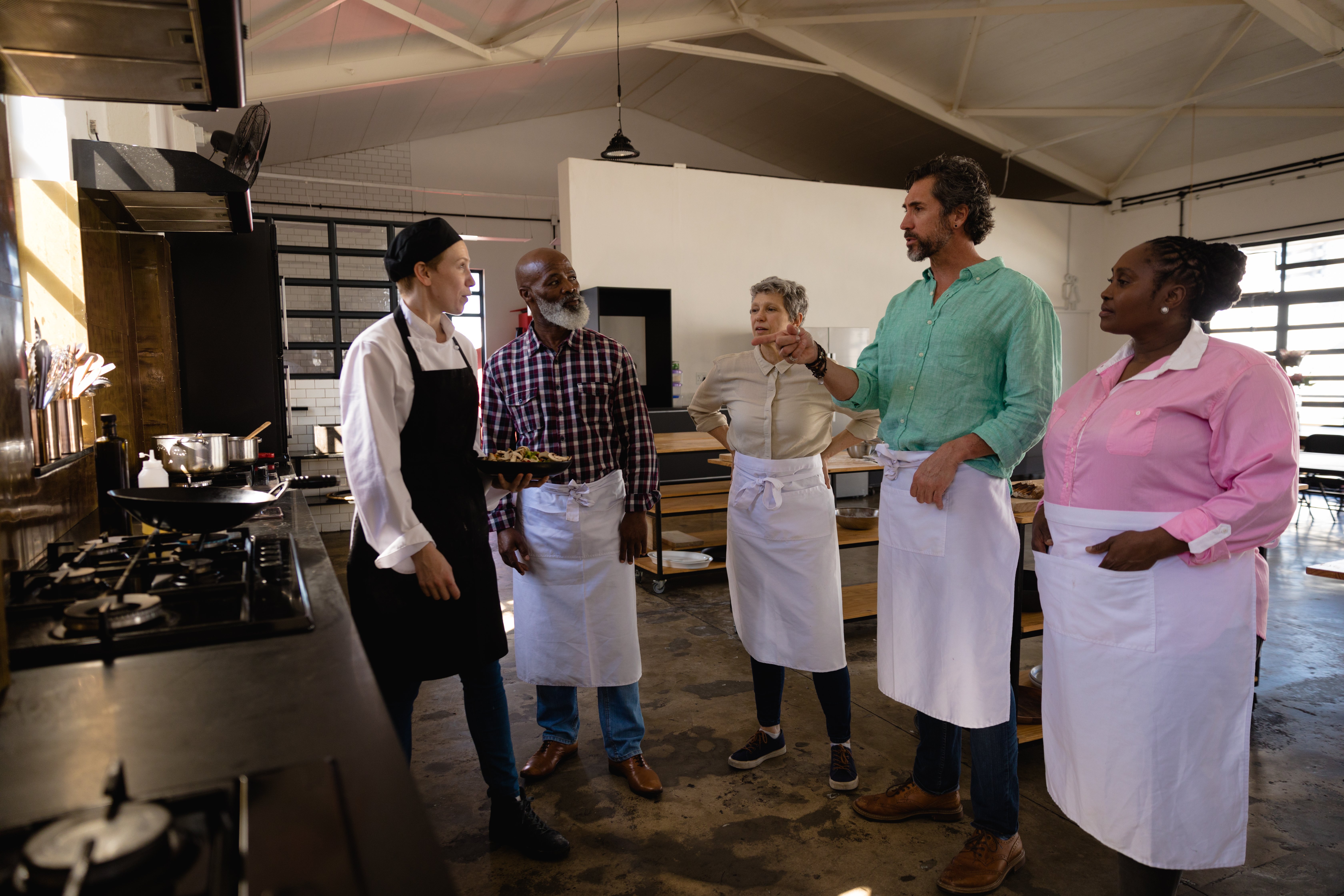 side-view-multi-ethnic-senior-group-adults-cookery-class-diverse-adult-students-listening-instructions-from-caucasian-female-chef-wearing-chefs-whites-black-hat-apron